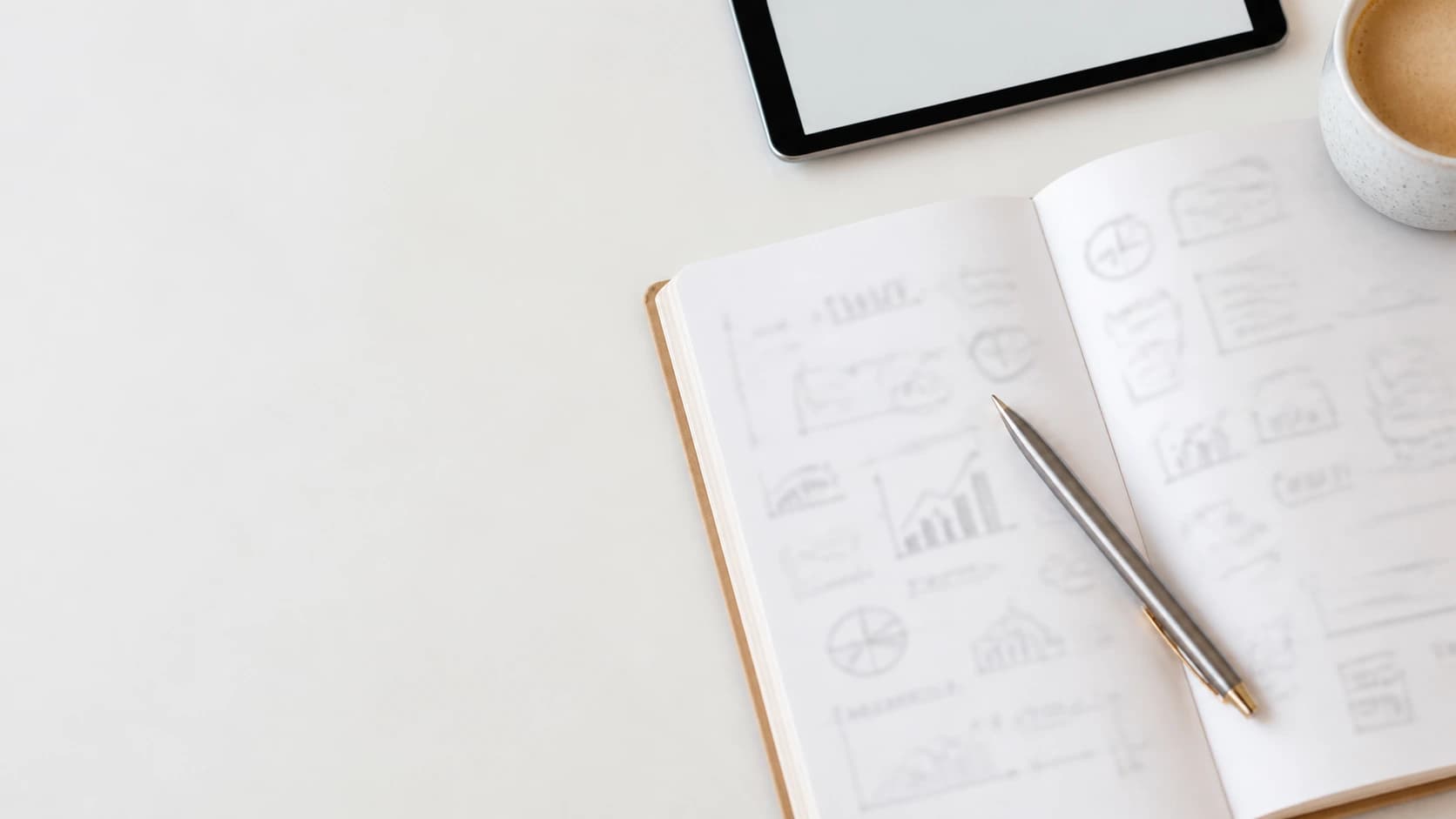 An open notebook of hand-drawn charts beside a tablet and a cup of coffee.