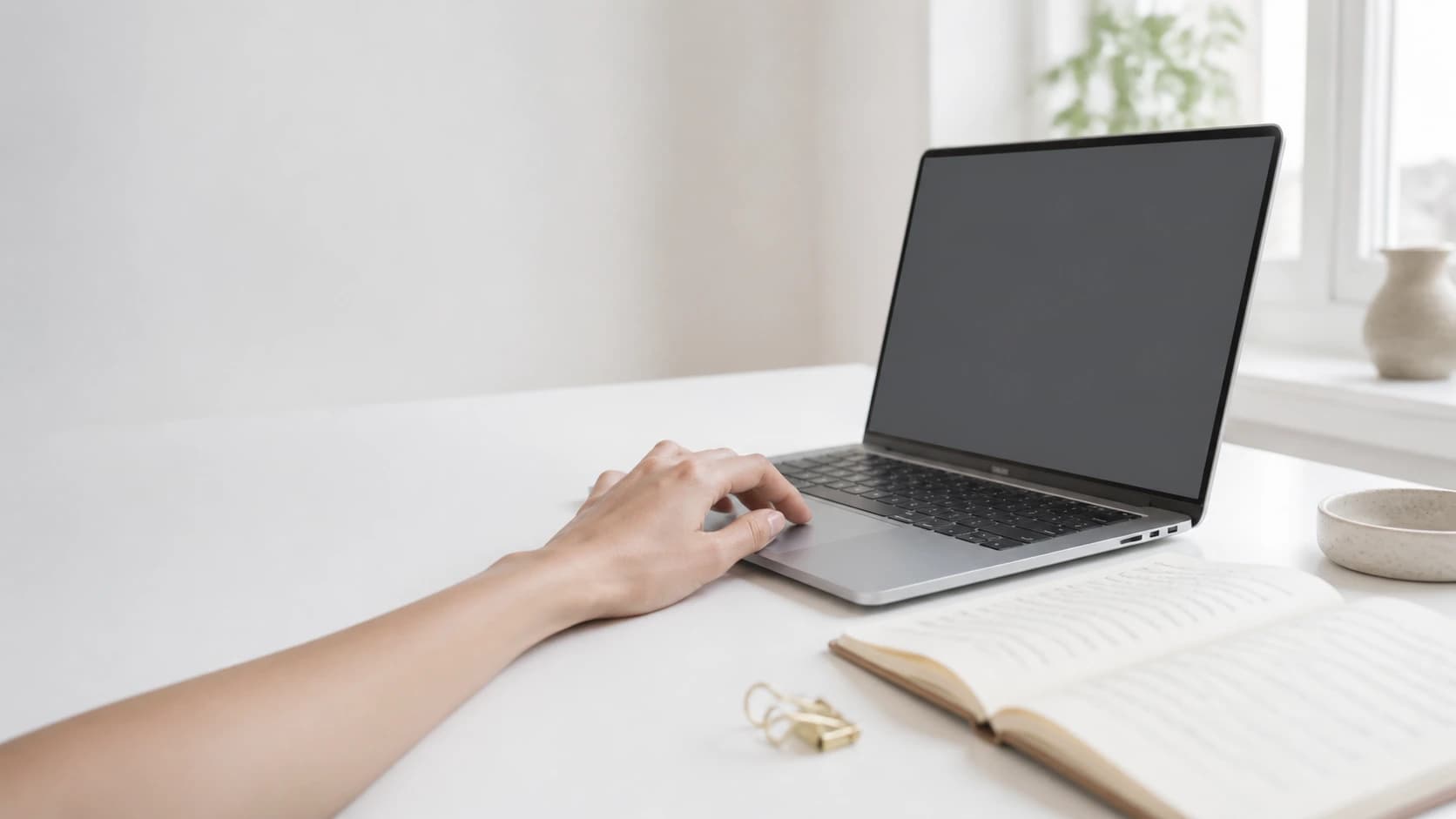 A hand resting on a laptop trackpad beside an open notebook on a bright white desk.